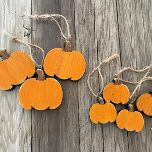 May include: Wooden pumpkin ornaments in various sizes, painted orange with brown stems. Each ornament has a twine loop for hanging. The pumpkins are arranged on a weathered, gray wooden surface, perfect for fall decor.