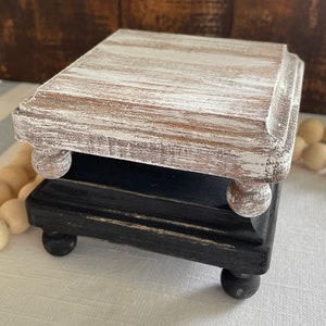 May include: A two-tiered wooden riser with a distressed white top tier and a black bottom tier. Both tiers have turned wooden legs.