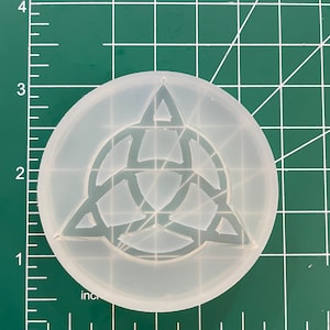 May include: A clear, circular silicone mould featuring a Celtic knot design. The mould is approximately 7.6 cm in diameter and sits on a green grid surface. The design is a series of interlocking shapes.