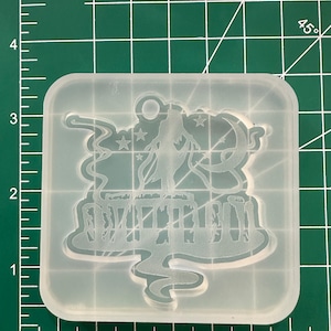 May include: A clear silicone mold with a rounded rectangular shape. The mold features a detailed design of a figure with stars, a crescent moon, and a cityscape. The mold is on a green cutting mat with inch measurements.
