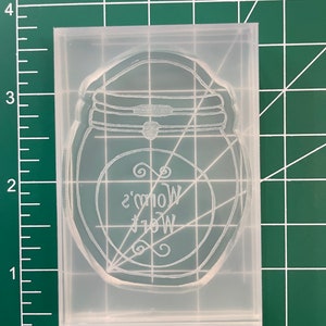 May include: A clear, rectangular mold with a jar design. The jar has a lid and a circular label with the text "Mom's Wet" in a decorative font. The mold is on a green cutting mat with inch measurements.