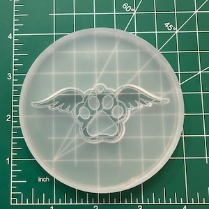 May include: A clear, circular silicone mold featuring a paw print design with angel wings. The mold is on a green cutting mat with inch measurements. The paw print is detailed with visible paw pad and claw impressions.