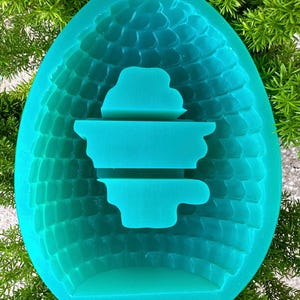 May include: A teal silicone mold in the shape of a pinecone. The mold has a textured exterior and a smooth interior with a layered design. The mold is surrounded by green foliage.