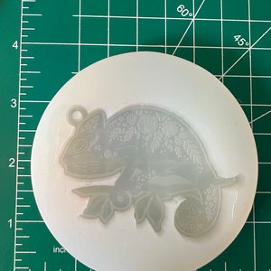 May include: A clear, circular silicone mold featuring a detailed chameleon design with floral accents. The chameleon is perched on a branch, and the mold includes a small loop for hanging. The mold is approximately 4 inches in diameter.