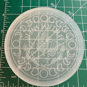 May include: A clear, circular mold with etched designs, including crescent moons, botanical elements, and the text "LAJITIAL MAGIC". The mold is approximately 4 inches in diameter and sits on a green cutting mat with inch measurements.