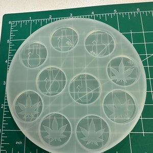 May include: A clear, circular silicone mold with multiple circular cavities. Each cavity features an etched design of either a bong or a cannabis leaf. The mold is on a green cutting mat with inch measurements.