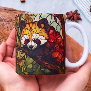 May include: White ceramic mug with a stained glass design of a red panda sitting on a branch. The panda is surrounded by leaves and flowers in shades of red, orange, yellow, and green.