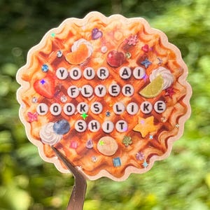 May include: A round, scalloped-edge sticker featuring a waffle illustration topped with fruit, whipped cream, and small colourful charms. The text "YOUR AI FLYER LOOKS LIKE SHIT" is printed in the centre using white circular beads with black letters.