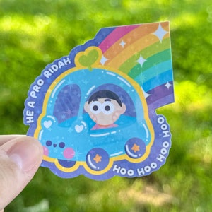 May include: A blue cartoon car with a rainbow and stars. The car has a smiling face and the text "HE A PRO RIDAH" and "HOO HOO HOO" around it. A cartoon character with brown hair is driving the car.