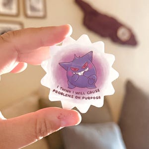 May include: A clear sticker with a purple cartoon character and the text "I THINK I WILL CAUSE PROBLEMS ON PURPOSE". The character has a mischievous grin and red eyes, set against a pink background.