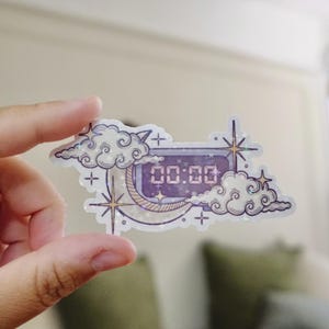 Puede incluir: Una pegatina holográfica con un reloj digital que muestra "00:00" en morado. El reloj está rodeado de nubes, estrellas y una luna creciente. La pegatina tiene un tema celestial y onírico.