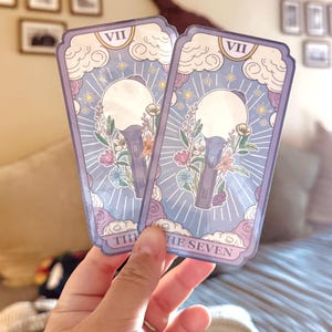 Puede incluir: Dos cartas de tarot con un diseño lila claro y azul, con un micrófono estilizado y elementos florales. Las cartas tienen un motivo de nubes y estrellas, con el texto "THE SEVEN" en la parte inferior y el número romano "VII" en la parte superior.