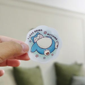 May include: A die-cut sticker featuring a cartoon Snorlax character in blue and white, lying down with the text "I LOVE DOING NOTHING". The sticker is surrounded by small pink flowers.