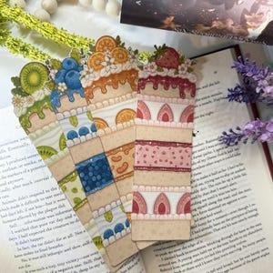 May include: Four decorative bookmarks with a cake design. Each bookmark features a different fruit topping: kiwi, blueberries, orange slices, and strawberries. The bookmarks are rectangular and made of paper.
