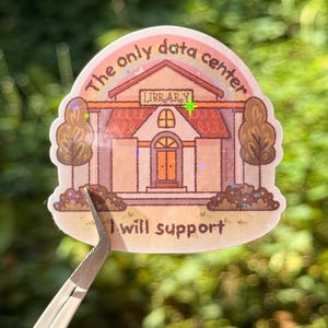 May include: A die-cut sticker featuring a light pink building with a sign that reads LIBRARY. Above the building is a rainbow and the text The only data center. Below the building is the text I will support. The sticker has a holographic finish.