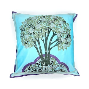 May include: Square throw pillow with a light blue background and a tree design. The tree has a dark brown trunk and branches, with green and white leaves and flowers. The pillow has a purple border.