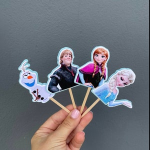 May include: Four Frozen themed cupcake toppers. The toppers feature Olaf, Elsa, Anna, and Kristoff. The toppers are printed on cardstock and attached to wooden sticks.