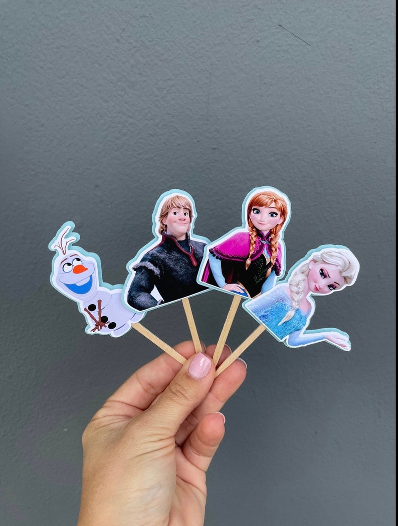 May include: Four Frozen themed cupcake toppers. The toppers feature Olaf, Elsa, Anna, and Kristoff. The toppers are printed on cardstock and attached to wooden sticks.
