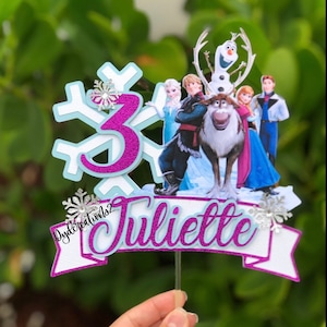 May include: A purple and white cake topper with a snowflake design, the number 3, and the name "Juliette". The topper features characters from the movie Frozen, including Olaf, Elsa, Anna, Kristoff, and Sven.