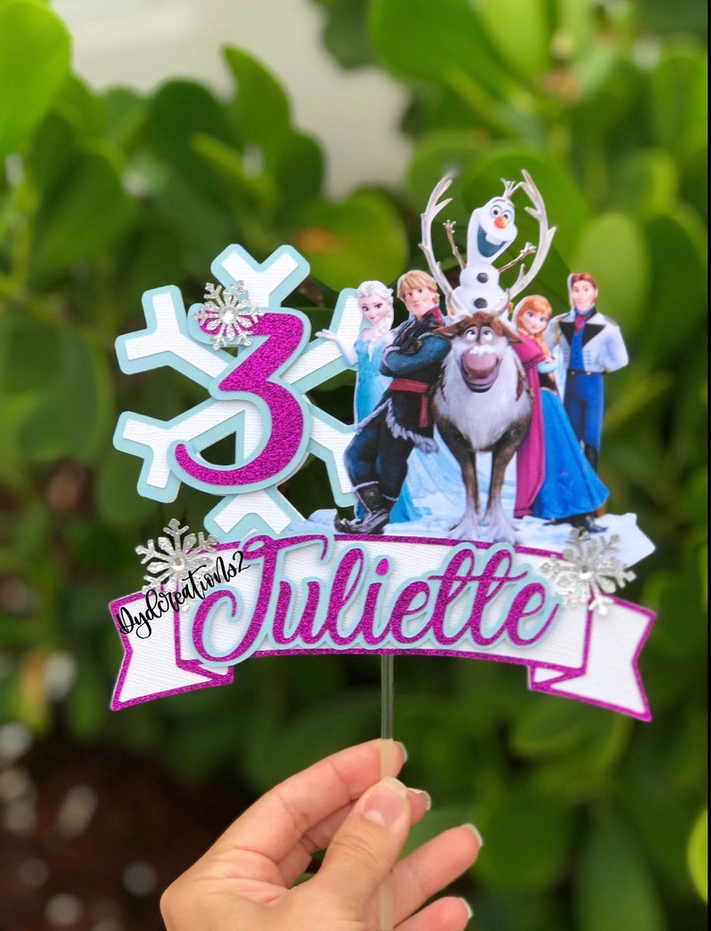 May include: A purple and white cake topper with a snowflake design, the number 3, and the name "Juliette". The topper features characters from the movie Frozen, including Olaf, Elsa, Anna, Kristoff, and Sven.
