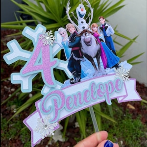 May include: A purple glitter number 4 cake topper with a blue and white snowflake design. The topper features characters from the movie Frozen, including Olaf, Kristoff, Sven, Anna, and Elsa. The topper also includes a blue and white banner with the name Penelope.