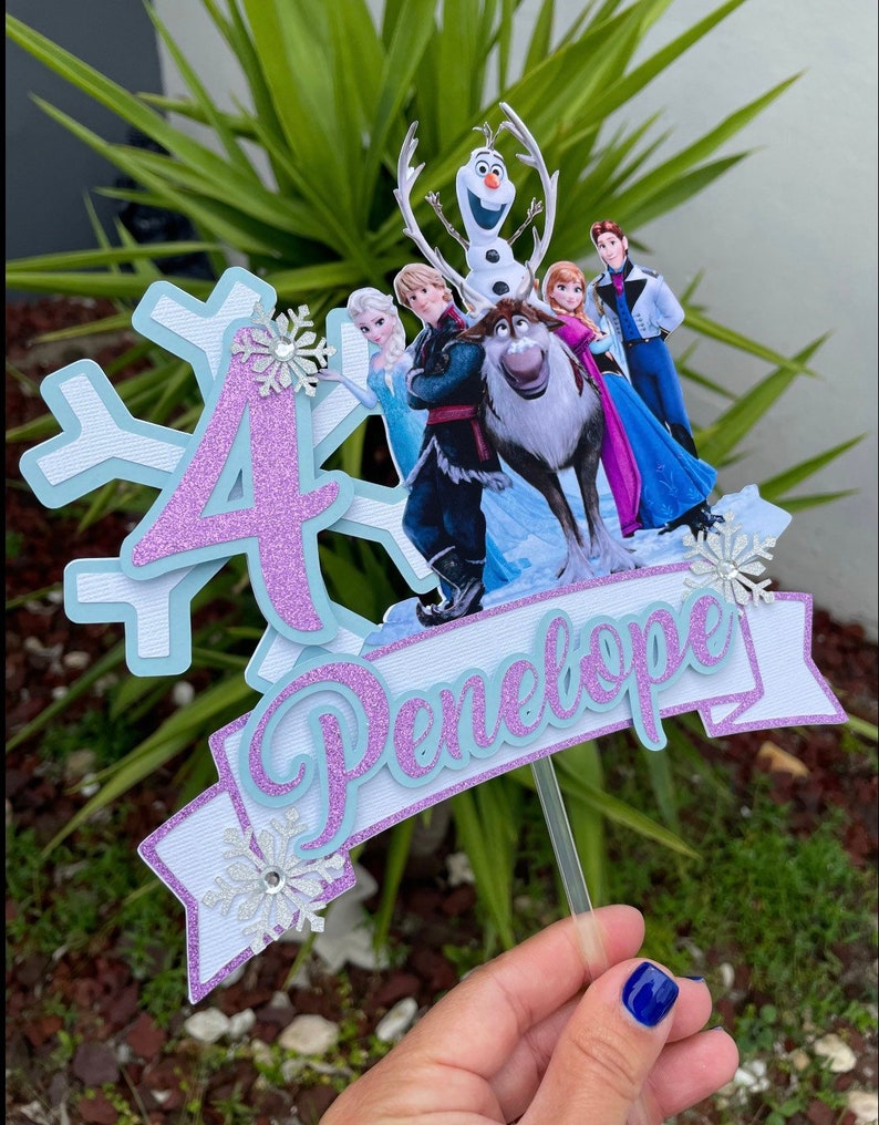 May include: A purple glitter number 4 cake topper with a blue and white snowflake design. The topper features characters from the movie Frozen, including Olaf, Kristoff, Sven, Anna, and Elsa. The topper also includes a blue and white banner with the name Penelope.
