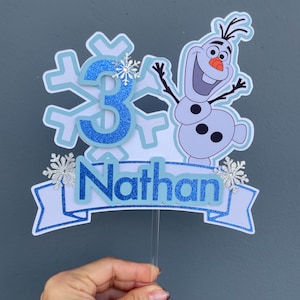May include: A cake topper featuring the number "3" in blue glitter, a snowflake design, and the character Olaf from Frozen. The name "Nathan" is displayed on a banner. The topper is held by a clear plastic stick.