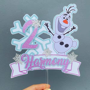 May include: A birthday cake topper featuring the number 2 in glittery pink, a cartoon image of Olaf the snowman, and the name "Harmony" in pink script. The design includes snowflakes and is mounted on a clear stick.