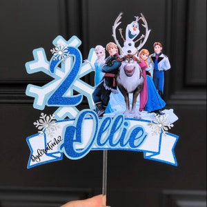May include: Blue and white cake topper with a snowflake, the number 2, and the name "Ollie" in blue glitter. The topper features characters from the movie Frozen, including Olaf, Elsa, Anna, Kristoff, and Sven.