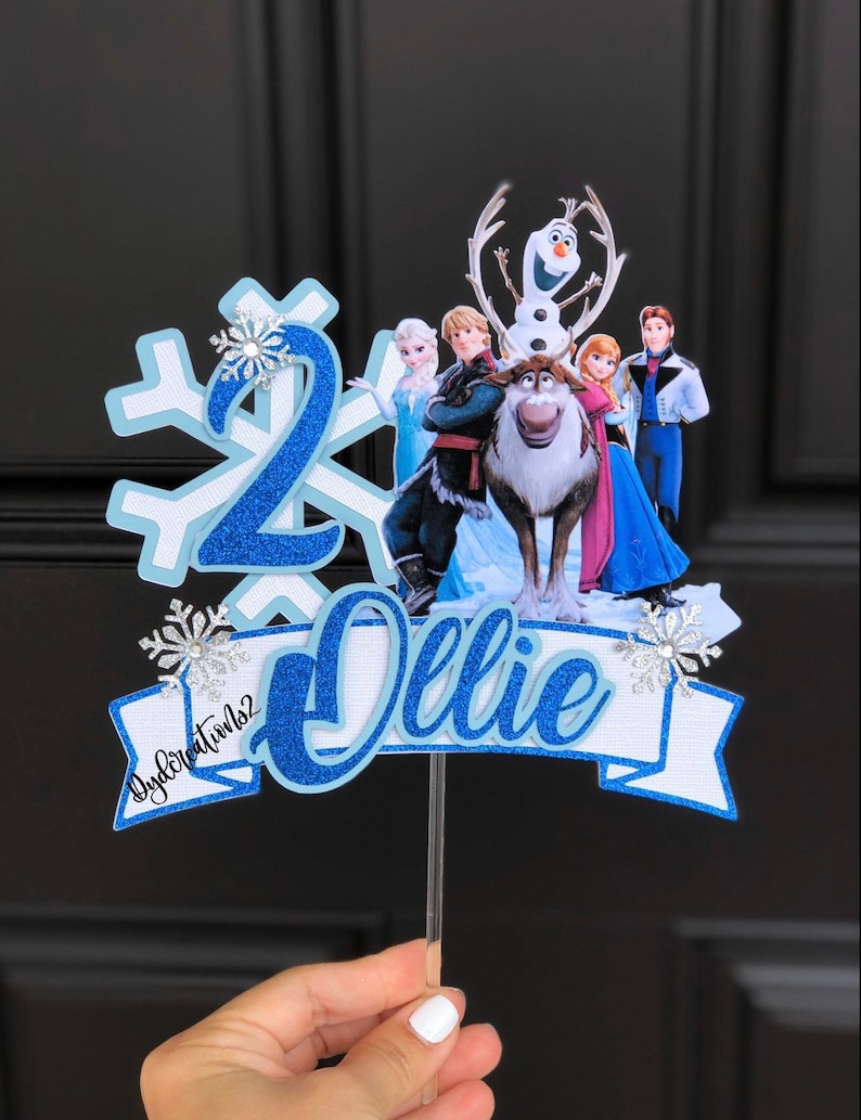 May include: Blue and white cake topper with a snowflake, the number 2, and the name "Ollie" in blue glitter. The topper features characters from the movie Frozen, including Olaf, Elsa, Anna, Kristoff, and Sven.