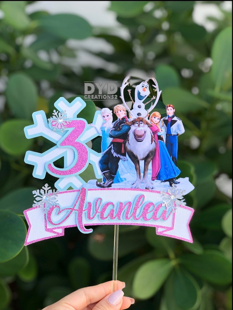 May include: A Frozen-themed cake topper featuring a pink number 3, a snowflake, and a group of Frozen characters including Olaf, Kristoff, Sven, Anna, and Elsa. The topper also includes a pink banner with the name "Avalea" written in blue.
