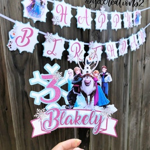 May include: A pink and blue Frozen themed cake topper with a snowflake, the number 3, and the name "Blakely". The topper features characters from the movie Frozen, including Elsa, Anna, Olaf, Kristoff, and Sven.