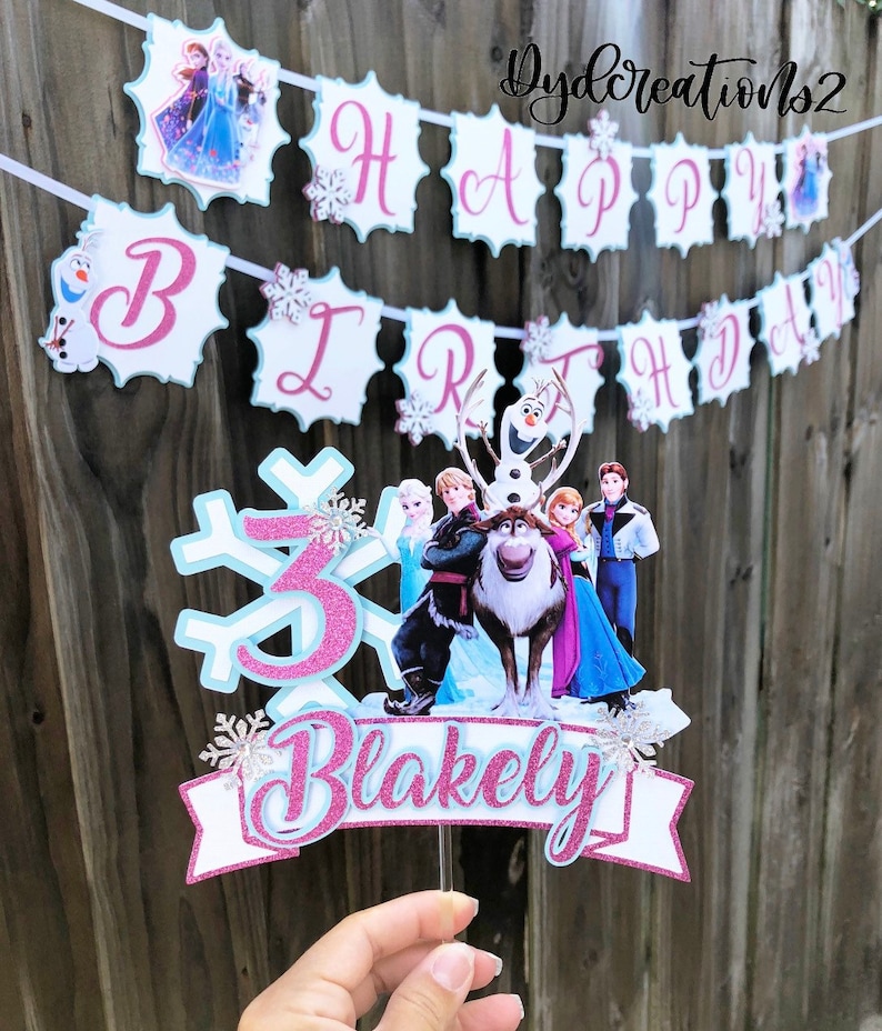 May include: A pink and blue Frozen themed cake topper with a snowflake, the number 3, and the name "Blakely". The topper features characters from the movie Frozen, including Elsa, Anna, Olaf, Kristoff, and Sven.