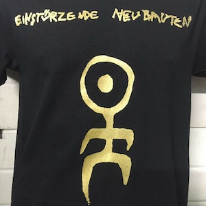 May include: Black t-shirt with a gold graphic of a stylised human figure and the text "EINSTÜRZENDE NEUBAUTEN" printed on the front.