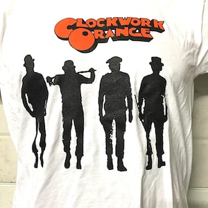 May include: White t-shirt featuring the text "Clockwork Orange" in orange and black. Below the text are four black silhouettes of figures in hats and suits, each holding a different object. The shirt is made of a soft, white material.