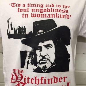 May include: White t-shirt with a black and red graphic. The design features a portrait of a man in a hat, the words "The Witchfinder General," and a quote in red lettering.
