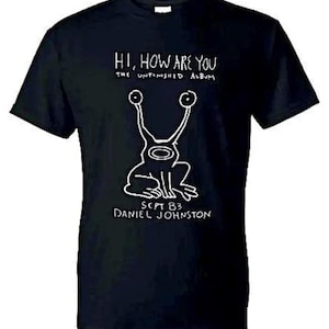 May include: Black t-shirt with a white drawing of a creature with large eyes and the text "HI, HOW ARE YOU" and "THE UNFINISHED ALBUM". The text "SEPT 83 DANIEL JOHNSTON" is below the illustration.