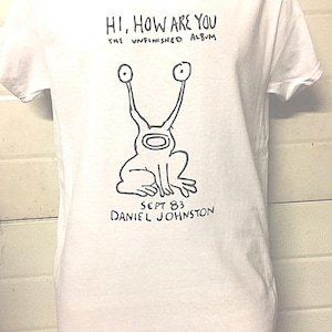 May include: White t-shirt featuring a black and white illustration of a creature with two eyes and long necks. The text "HI, HOW ARE YOU THE UNFINISHED ALBUM SEPT 83 DANIEL JOHNSTON" is printed on the shirt.