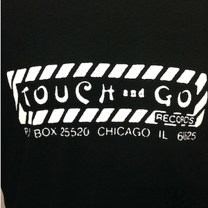 May include: Black t-shirt with a white graphic of the text "TOUCH and GO RECORDS" with the address "P.O. BOX 25520 CHICAGO IL 60625" underneath.