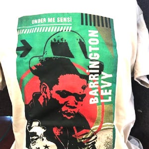 May include: Off-white t-shirt featuring a graphic of Barrington Levy in red and black, with a green and red background. The text "UNDER ME SENSI" and "BARRINGTON LEVY" are also visible. The shirt has a crew neck and short sleeves.