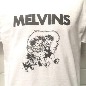 May include: White t-shirt with a black graphic of two children and a dog. The text "MELVINS" is printed in black at the top of the shirt.