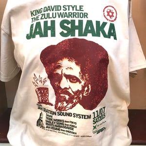 May include: Cream-colored t-shirt featuring a maroon portrait of a man with a beard and the words "JAH SHAKA" in large green letters. The shirt also includes text about a sound system and a tour date.