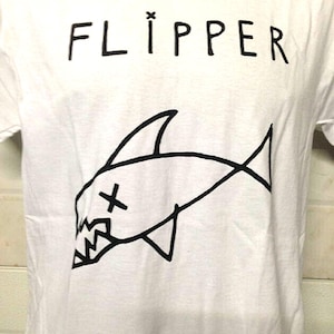 May include: White t-shirt with black text that reads "FLIPPER" and a black cartoon drawing of a shark with a cross on its head.