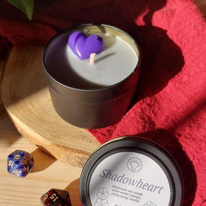 Könnte beinhalten: Eine schwarze Zinnkerze mit einem herzförmigen Wachsdesign in Lila. Die Kerze steht auf einer Holzoberfläche neben einem roten Tuch und zwei Würfeln. Auf dem Etikett der Kerze steht "Shadowheart" und die Duftnoten.