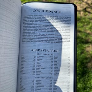 Pode incluir: B&iacute;blia aberta com as palavras "CONCORDANCE" e "ABBREVIATIONS". As p&aacute;ginas exibem listas de termos e refer&ecirc;ncias b&iacute;blicas. A B&iacute;blia tem uma capa preta e est&aacute; aberta na grama.
