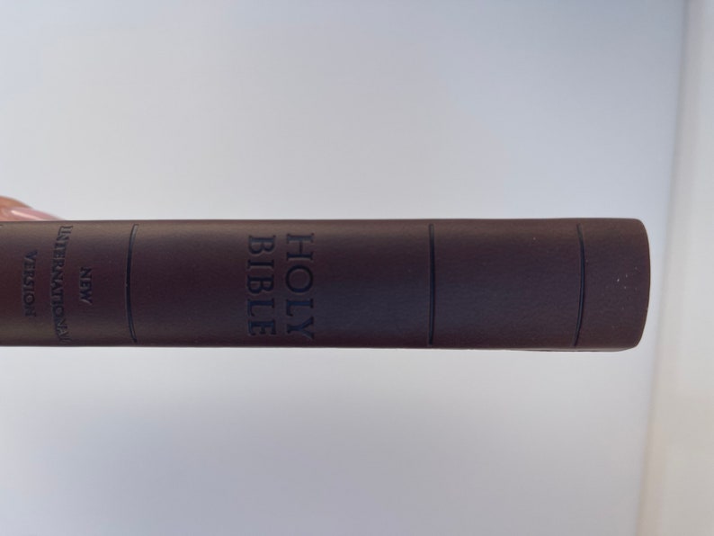 K&ouml;nnte beinhalten: Eine braune, ledergebundene Heilige Bibel mit den Worten "HOLY BIBLE" auf dem R&uuml;cken gepr&auml;gt. Der Text "NEW INTERNATIONAL VERSION" ist seitlich aufgedruckt.