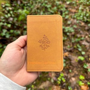 May include: A small, tan-colored book with rounded corners. The cover features a floral design and the embossed text "FirstName LastName." The book is held against a blurred green background.