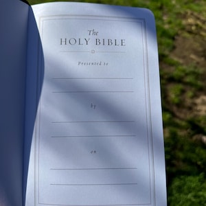 Pode incluir: B&iacute;blia aberta com o t&iacute;tulo "The Holy Bible" na p&aacute;gina interior. A p&aacute;gina tem uma borda decorativa e linhas para o nome do destinat&aacute;rio e a data. O fundo est&aacute; desfocado com luz natural.
