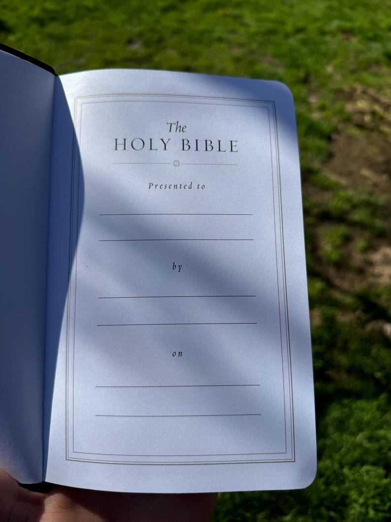 Pode incluir: B&iacute;blia aberta com o t&iacute;tulo "The Holy Bible" na p&aacute;gina interior. A p&aacute;gina tem uma borda decorativa e linhas para o nome do destinat&aacute;rio e a data. O fundo est&aacute; desfocado com luz natural.