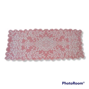 Può includere: Un runner da tavolo in crochet bianco con un bordo a festoni e un motivo floreale. Il runner è su uno sfondo rosso.